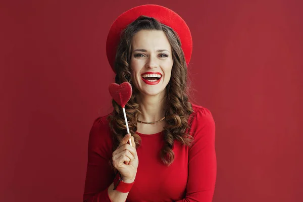 ハッピーバレンタイン 笑顔現代的な中年女性で赤いドレスとベレー帽に赤い背景に対してハート型のキャンディーオンスティック — ストック写真