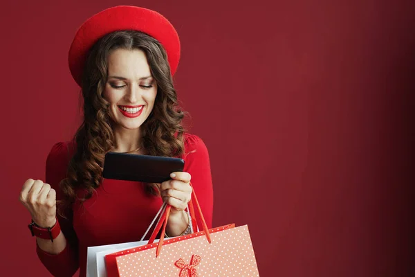 ハッピーバレンタイン 赤いドレスとベレー帽の長い波状の髪を持つ幸せなエレガントな女性は スマートフォンのアプリケーションを使用してショッピングバッグと赤の背景にあり 電子商取引のウェブサイト上でオンライン購入を行う — ストック写真