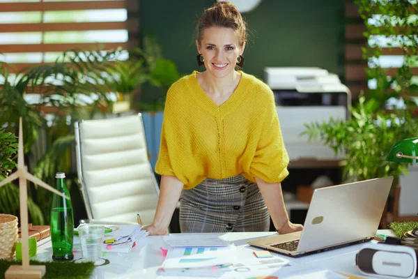 エレガントな笑顔40歳の小さなビジネスの所有者女性で黄色のセーターでラップトップ作業でドキュメントで現代の緑のオフィス — ストック写真