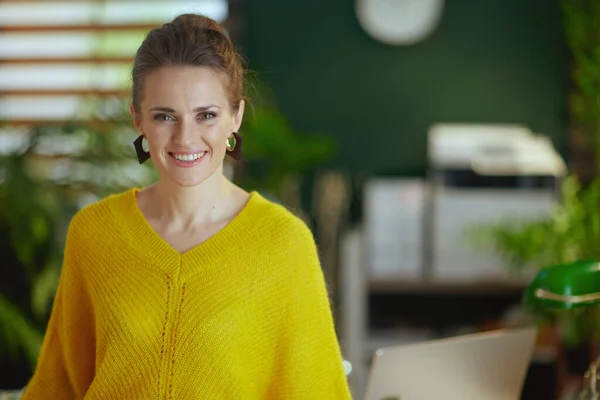 現代の緑のオフィスに立つ黄色のセーターに身を包んだ若い中小企業経営者の女性を笑顔 — ストック写真