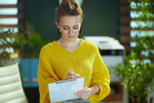 現代的な中年中小企業の所有者女性で黄色のセーターでクリップボードで現代的な緑のオフィス — ストック写真