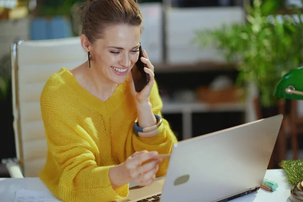 エレガントな中小企業経営者の女性を笑顔黄色のセーターにラップトップで話す現代の緑のオフィスのスマートフォン — ストック写真