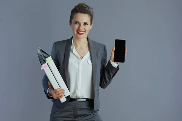 スマートフォンのブランク画面を示すフォルダとグレーのスーツで現代的な中年中小企業の所有者の女性を笑顔灰色の背景に隔離 — ストック写真