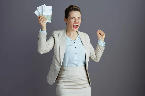 幸せなエレガントな小さなビジネスの所有者の女性で軽いビジネススーツでユーロマネーパック灰色の背景に対して上昇腕喜び — ストック写真