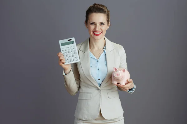 快乐的现代小企业主女人穿着轻便的西装 带着小猪银行和灰色背景的计算器 — 图库照片