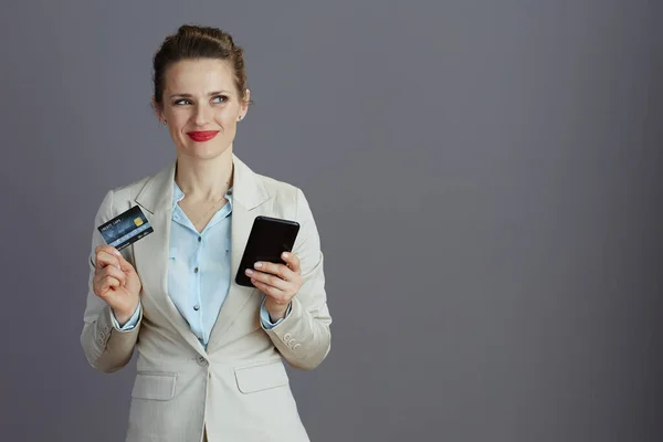 優雅な女性社員を笑顔にするライトビジネススーツスマートフォンとクレジットカードでグレーに隔離された — ストック写真