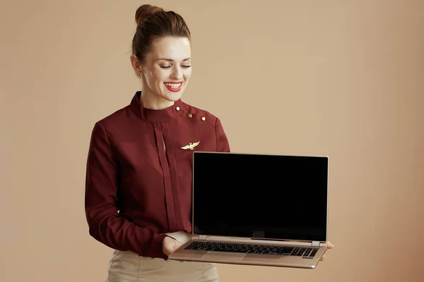 smiling modern female air hostess on beige background showing laptop blank screen.