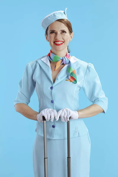 smiling elegant flight attendant woman against blue background in blue uniform with travel bag looking up at copy space.