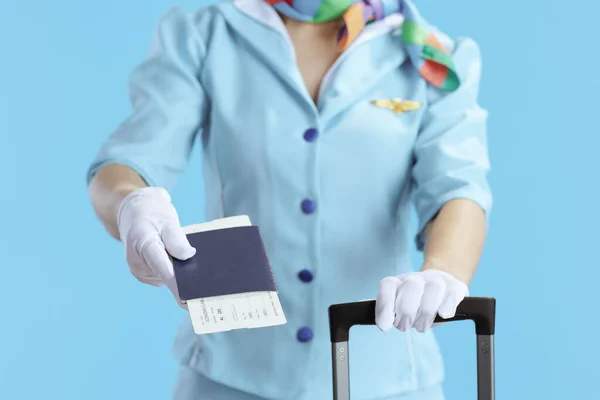 Closeup on stylish female flight attendant isolated on blue background in blue uniform with flight tickets, passport and wheel bag.