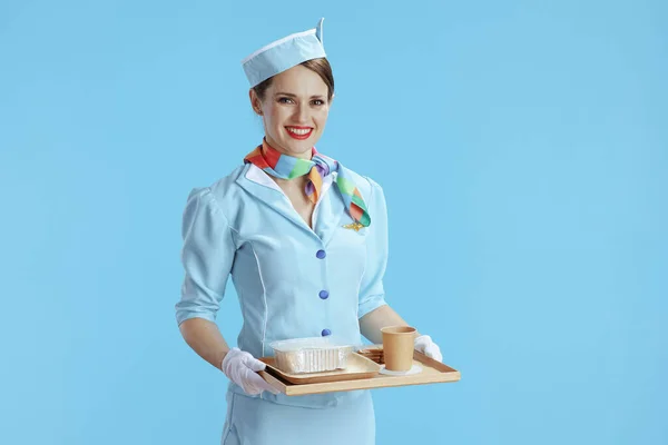 在蓝色背景下 穿着蓝色制服 端着一盘食物的现代空姐微笑着 — 图库照片