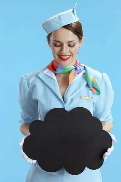 smiling elegant female air hostess on blue background in blue uniform showing blank cloud shape board.