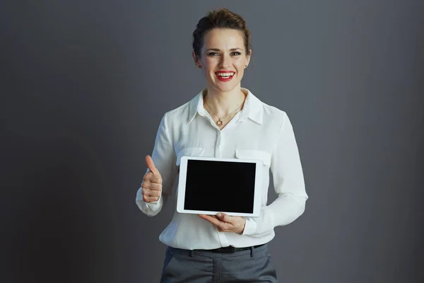 笑顔スタイリッシュな小さなビジネス所有者女性で白いブラウスショータブレットPc空白の画面と親指アップに対してグレーの背景 — ストック写真