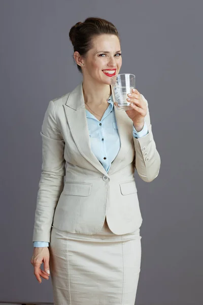 穿着浅色西服 杯子里的水与灰色隔离的现代小企业主女人微笑着 — 图库照片