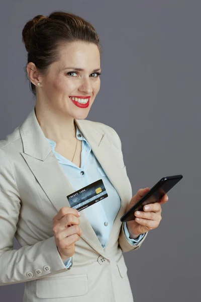 スマートフォンやクレジットカードをグレーの背景に孤立させたライトビジネススーツを着た優雅な女性労働者の笑顔 — ストック写真