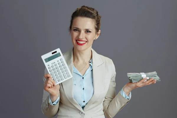 幸せなスタイリッシュな中年女性労働者でライトビジネススーツで計算機とドルのお金パックでグレーに隔離 — ストック写真