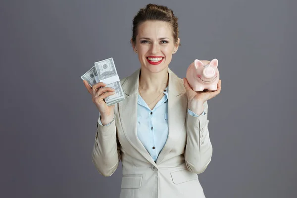 現代の40歳のビジネス女性を笑顔でピンクの銀行とドルのお金パックで軽いビジネススーツグレーに隔離された — ストック写真