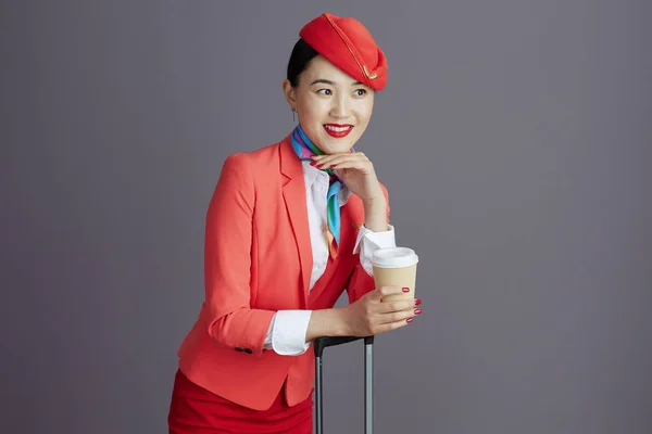 smiling elegant asian female air hostess in red skirt, jacket and hat uniform with coffee cup and trolley bag looking at copy space against grey background.