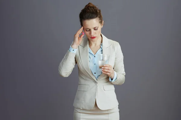 stressed modern 40 years old woman worker in a light business suit with glass of water and pill isolated on grey.