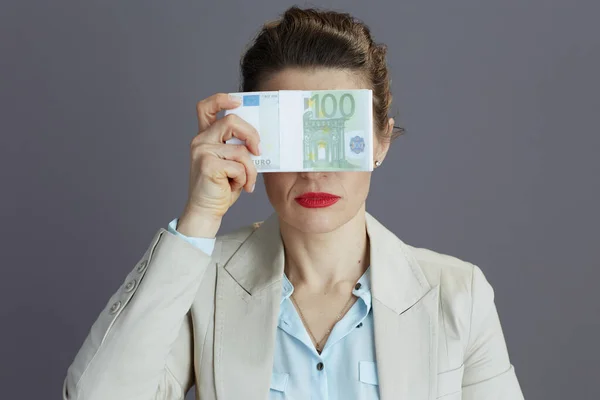 trendy 40 years old business woman in a light business suit with euros money packs showing see no evil gesture isolated on gray background.