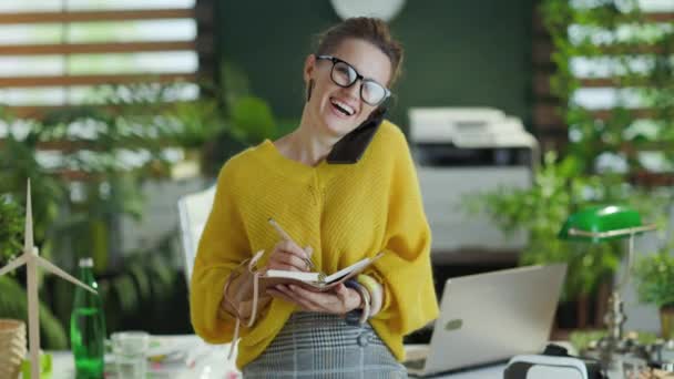 幸せな流行の小さなビジネスの所有者女性で黄色のセーターノートブックは現代の緑のオフィスでスマートフォン上で話す — ストック動画