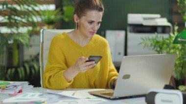 Modern yeşil ofiste akıllı telefon ve dizüstü bilgisayarı olan sarı kazaklı, modern küçük işletme sahibi kadın..