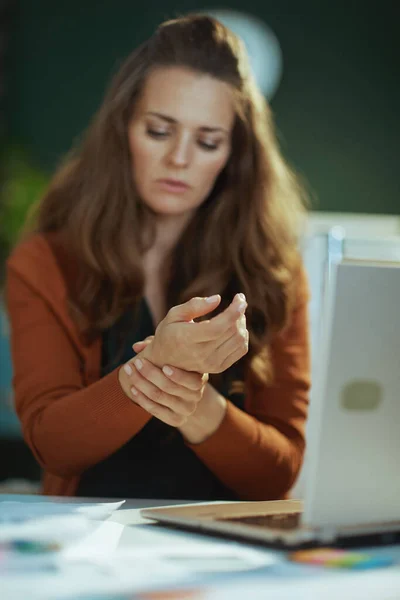 中年妇女小企业主穿着便携笔记本电脑和手腕在绿色办公室里痛苦不堪的衣服 — 图库照片
