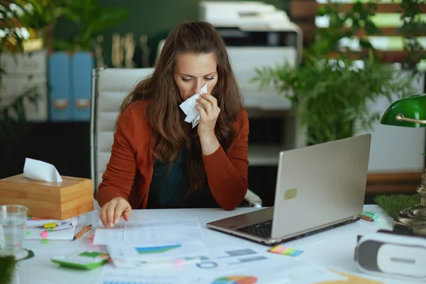 ラップトップとナプキンを持つスタイリッシュな中小企業の所有者の女性が鼻を吹いて 現代の緑のオフィスで文書を扱う — ストック写真
