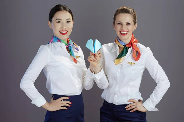 smiling modern flight attendant women in blue skirt, white shirt and scarf with paper airplane against gray background.