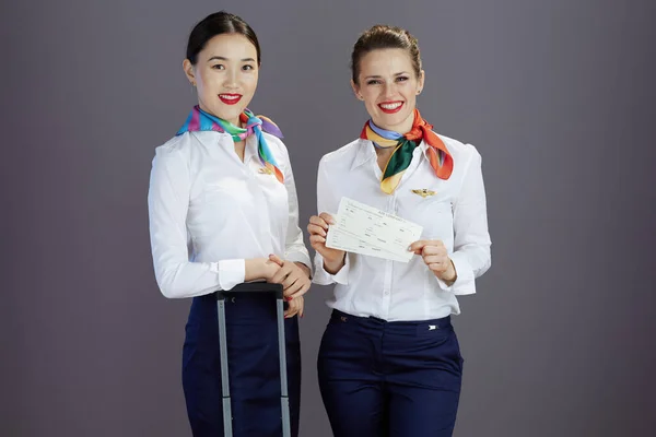 happy elegant air hostess women in blue skirt, white shirt and scarf with flight tickets and trolley bag against grey background.