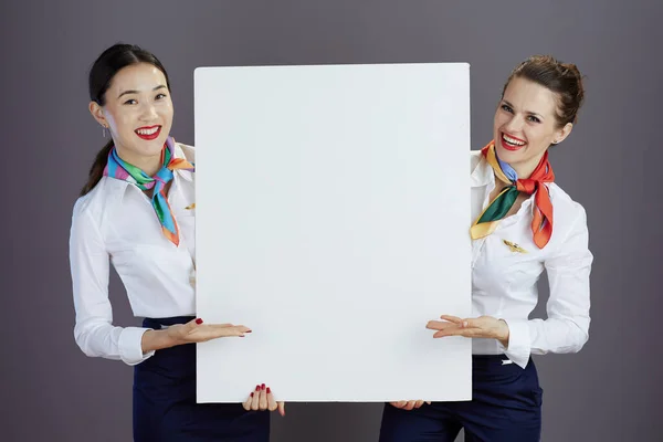 smiling elegant female flight attendants in blue skirt, white shirt and scarf with blank billboard isolated on grey.