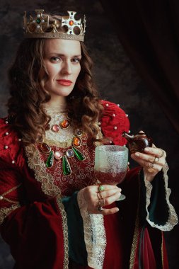 Kırmızı elbiseli ortaçağ kraliçesi koyu gri arka planda kadehe zehir döktü..