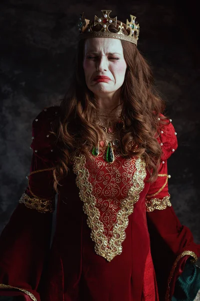 sad medieval queen in red dress with white makeup and crown on dark gray background.