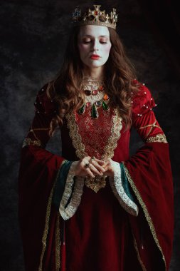 Kırmızı elbiseli, beyaz makyajlı ve tacı koyu gri arka planda olan ortaçağ kraliçesi..