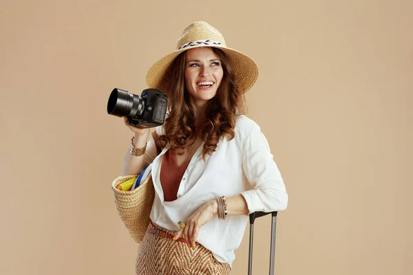 ビーチ休暇 白のブラウスとショートパンツで幸せなスタイリッシュな女性は 麦藁バッグ トロリーバッグ 写真カメラと藁帽子でベージュの背景に隔離されました — ストック写真