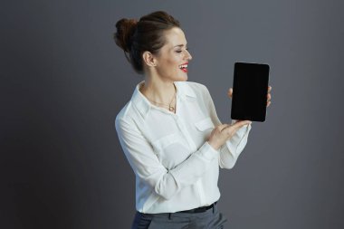 Beyaz bluz içinde gülümseyen zarif kadın çalışanı gri arkaplanda izole edilmiş tablet PC boş ekran gösteriyor.