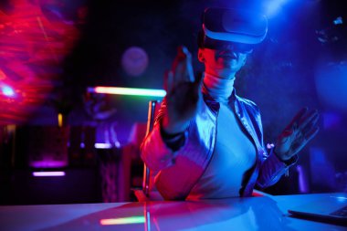 Neon metaevren fütüristik konsepti. Modern ofiste sanal gerçeklik gözlüklü meraklı orta yaşlı kadın..