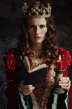 Kırmızı elbiseli, kitap, mum ve tacı koyu gri arka planda olan ortaçağ kraliçesi.