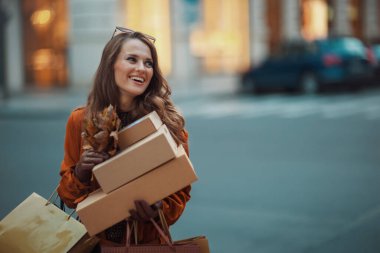 Merhaba Eylül. Gülümseyen 40 yaşındaki kahverengi trençkotlu kadın paketler, alışveriş torbaları ve sonbahar sarısı yapraklarıyla şehirde..