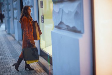 Merhaba sonbahar. Şehir merkezinde, alışveriş torbaları ve sonbahar sarısı yapraklarıyla turuncu trençkotlu modern 40 yaşındaki kadının tam boy portresi..