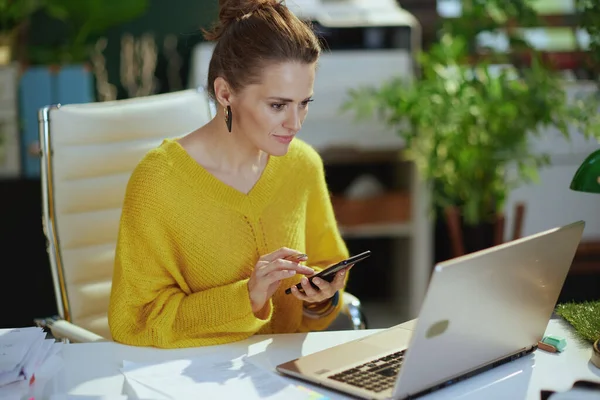 現代の緑のオフィスでスマートフォンを使用してノートパソコンと黄色のセーターで高価なトレンディーな中年中小企業の所有者の女性 — ストック写真