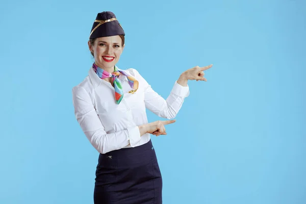 smiling elegant air hostess woman on blue background in uniform pointing at something.