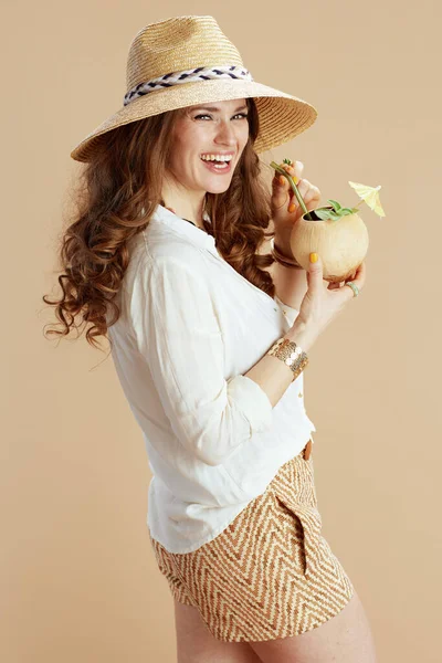 ビーチ休暇 白いブラウスでエレガントな主婦の笑顔とココナッツカクテルと麦藁帽子でベージュの背景に隔離されたショートパンツ — ストック写真