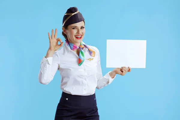 在蓝色背景下 穿着制服的现代女空姐微笑着 露出空白的纸片和好的手势 — 图库照片