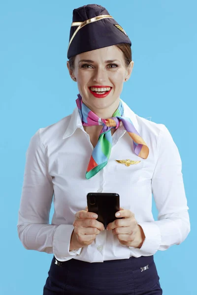 smiling modern air hostess woman against blue background in uniform sending text message using smartphone.