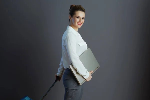 smiling 40 years old business woman in white blouse with laptop and trolley bag against gray background.