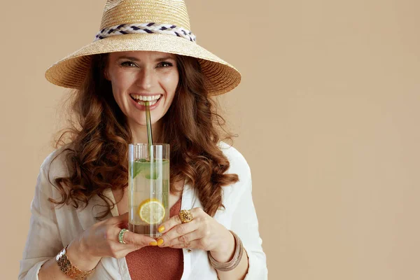 ビーチ休暇 エレガントな笑顔40歳の女性で白いブラウスとショートパンツでベージュの背景にカクテルと麦藁帽子 — ストック写真