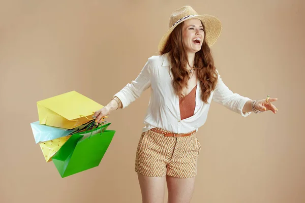 ビーチ休暇 現代的な笑顔40歳の女性で白ブラウスとショートパンツに対してベージュの背景でショッピングバッグとストロー帽子 — ストック写真