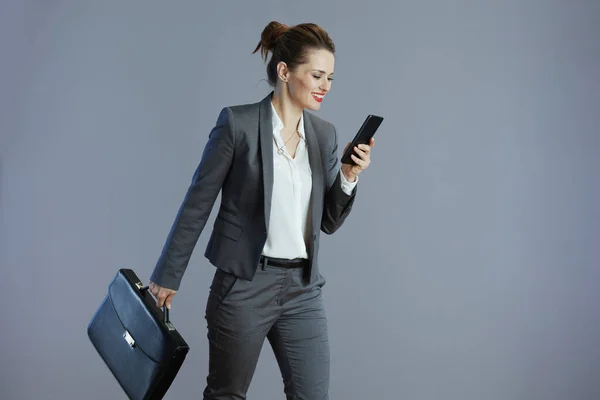 happy stylish female worker in gray suit with smartphone and briefcase isolated on gray background.