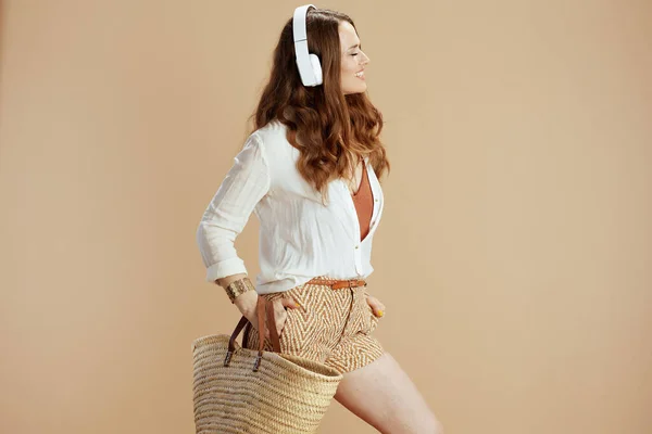 海滩度假 快乐的现代女性 40岁 身穿白色衬衫和短裤 背景为米黄色 头戴草袋和耳机 — 图库照片