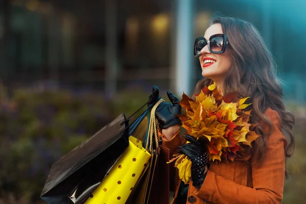 幸せな現代40歳の女性でオレンジトレンチコートでショッピングバッグと秋の黄色の葉近く店で市内 — ストック写真